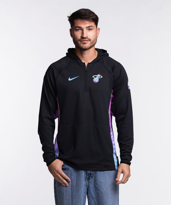 A man faces forward in a Nike Miami HEAT Original Vice Nights 1/2 Zip Hoodie, featuring blue and pink accents, paired with blue jeans. He has a neutral expression and stands against a plain white background.