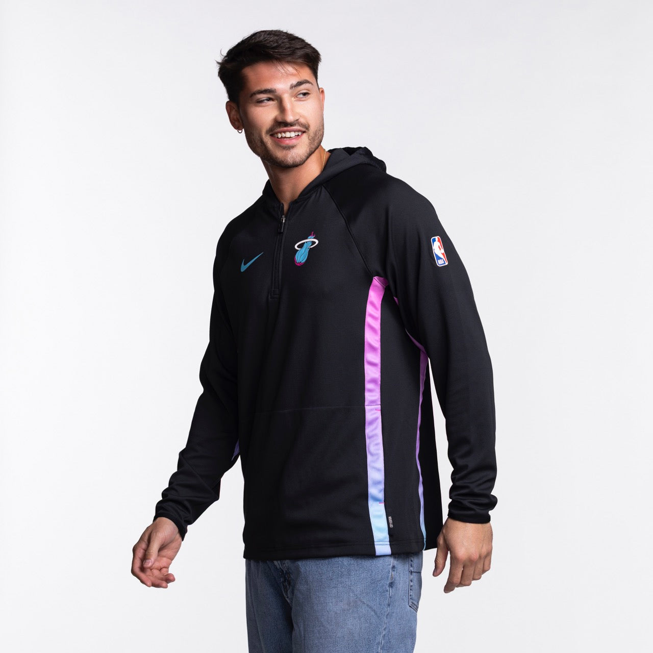 A man wears a Nike Miami HEAT Original Vice Nights 1/2 Zip Hoodie in black with teal and pink stripes, a light blue Nike logo, and light blue jeans, smiling against a plain white background.