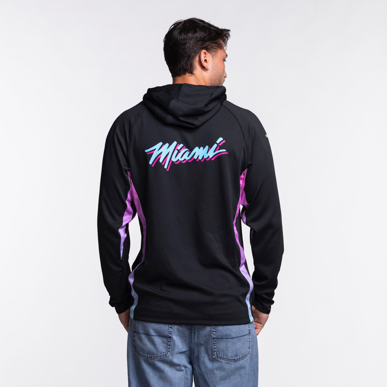 A person faces away, wearing a Nike Miami HEAT Original Vice Nights 1/2 Zip Hoodie in black with pink and blue “Miami” text on the back, paired with light blue jeans. The background is plain white.