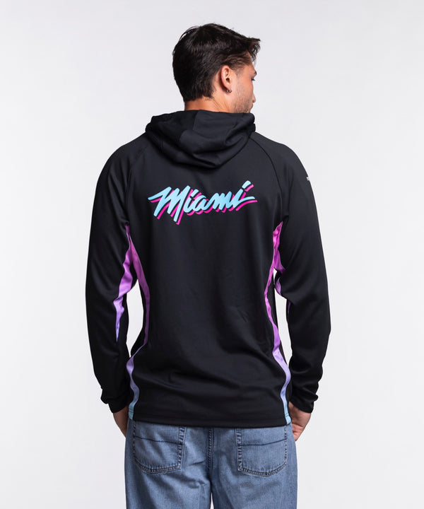 A man faces forward in a Nike Miami HEAT Original Vice Nights 1/2 Zip Hoodie, featuring blue and pink accents, paired with blue jeans. He has a neutral expression and stands against a plain white background.