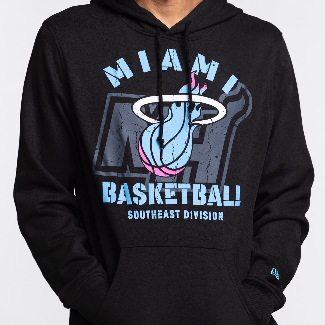 A person wears the New Era Miami HEAT Original Vice Nights Hoodie, a black standard fit, featuring a stylized Miami Heat logo and SOUTHEAST DIVISION text in blue and white. Machine washable for easy care.