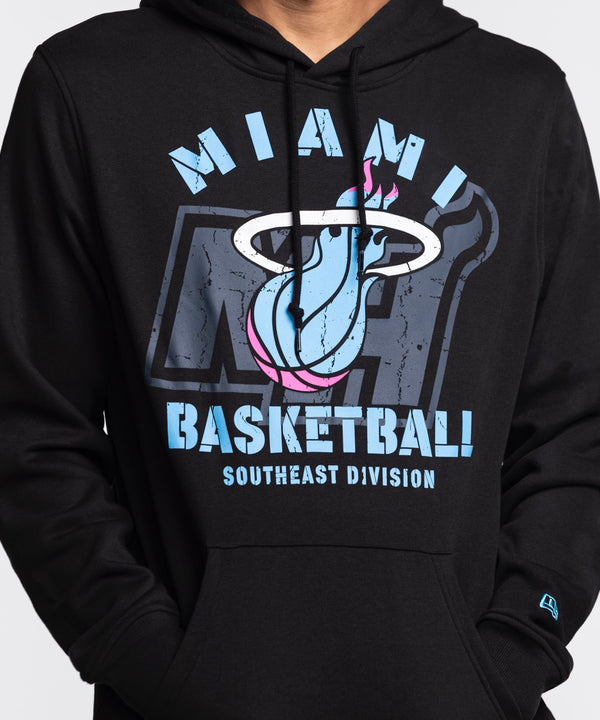 A man stands against a white background wearing the New Era Miami HEAT Original Vice Nights Logo Hoodie in black, paired with light blue jeans and a standard fit cotton-polyester t-shirt, hands in his pockets and looking to his left.