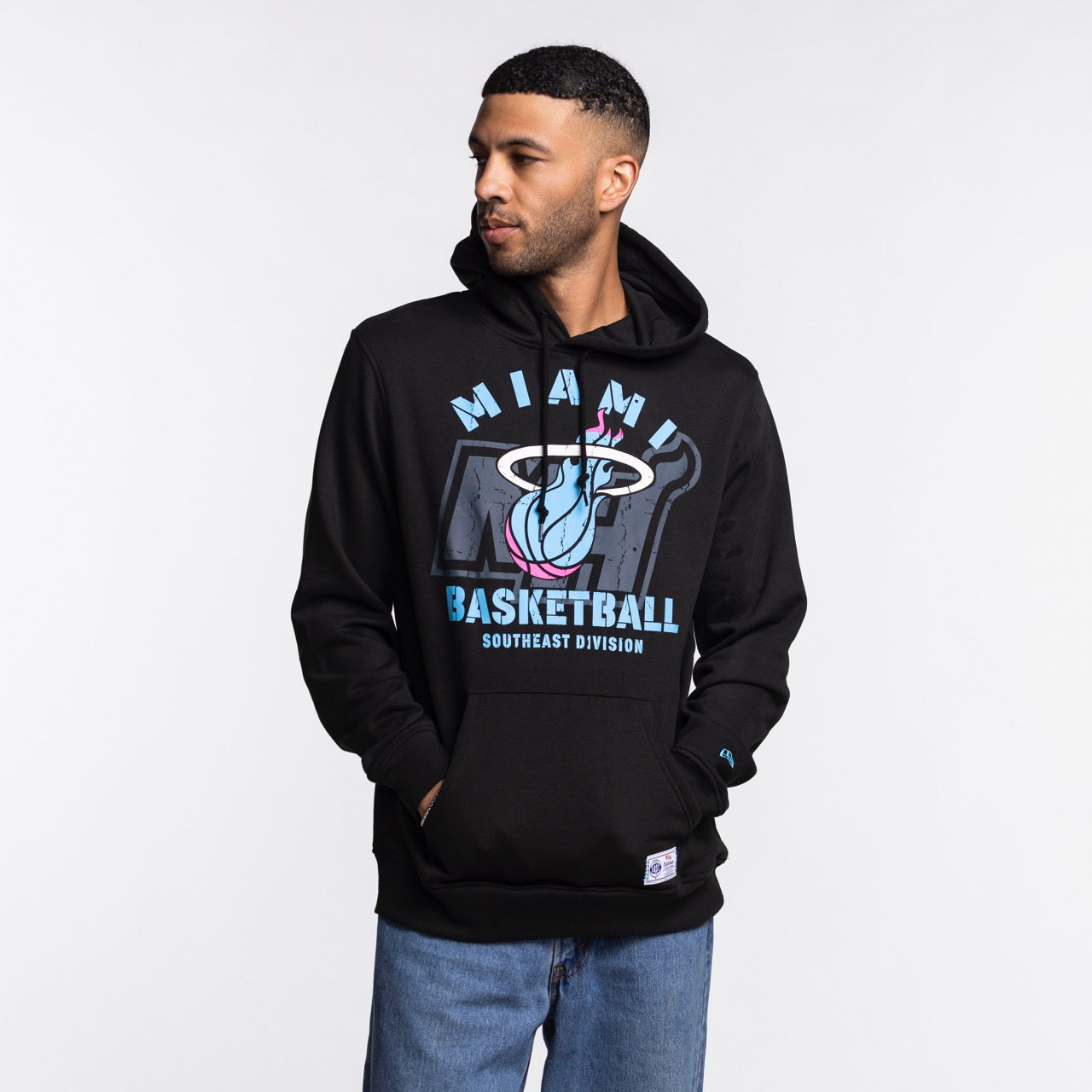 A man stands against a white background wearing the New Era Miami HEAT Original Vice Nights Logo Hoodie in black, paired with light blue jeans and a standard fit cotton-polyester t-shirt, hands in his pockets and looking to his left.