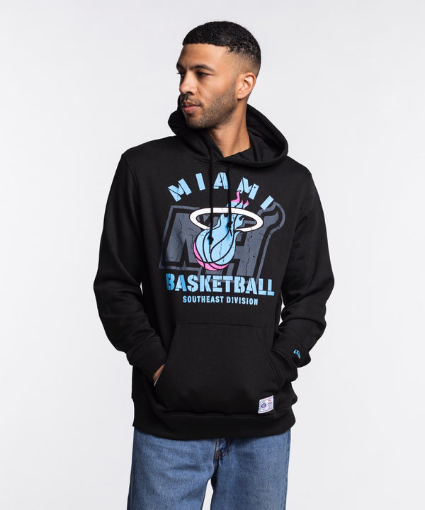 A man stands against a white background wearing the New Era Miami HEAT Original Vice Nights Logo Hoodie in black, paired with light blue jeans and a standard fit cotton-polyester t-shirt, hands in his pockets and looking to his left.