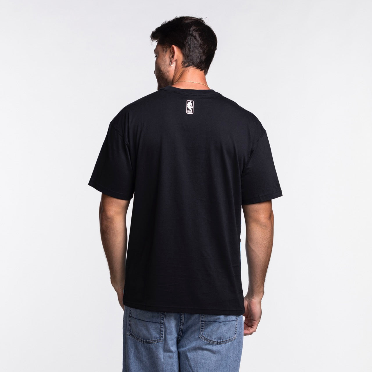 A man with short dark hair stands with his back to the camera, wearing a loose fit Nike Miami HEAT Original Vice Nights Big Logo Tee and light blue jeans against a white background.