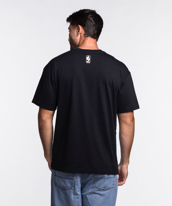 A person wearing the Nike Miami HEAT Original Vice Nights Big Logo Tee, a loose fit light blue Nike shirt with black sleeves and an abstract pink, black, and white graphic, stands against a plain white background.