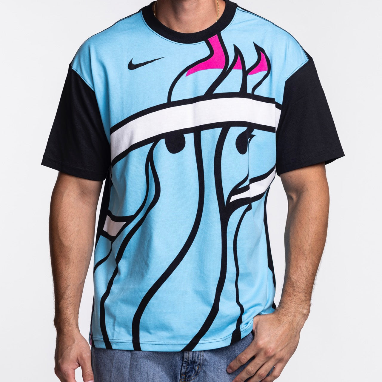 A person wearing the Nike Miami HEAT Original Vice Nights Big Logo Tee, a loose fit light blue Nike shirt with black sleeves and an abstract pink, black, and white graphic, stands against a plain white background.