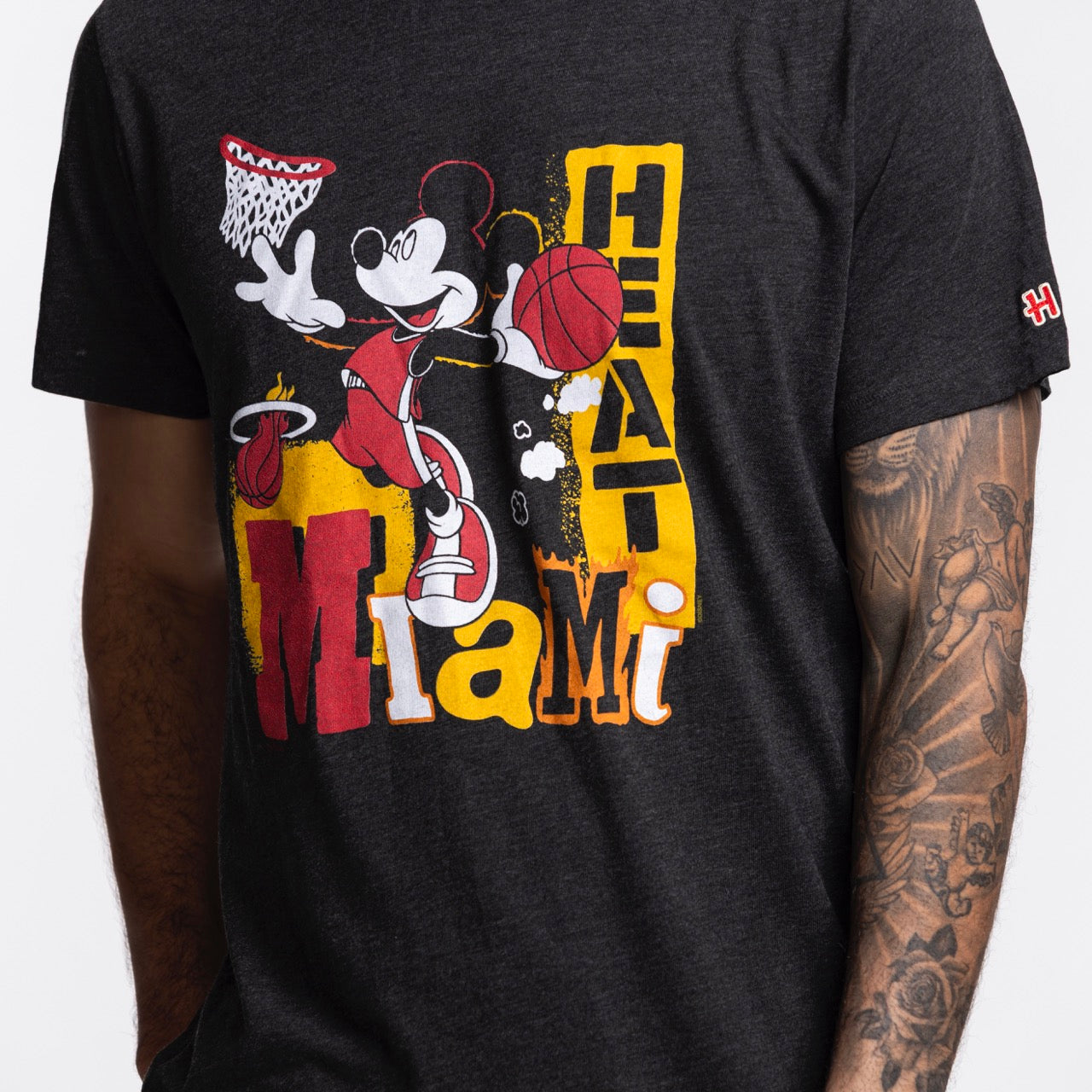 Someone wears the Homage Miami HEAT Mickey Basketball Tee, a black, standard fit T-shirt by Homage featuring Mickey Mouse playing basketball and bold Miami Heat text. Made from a soft polyester-cotton blend. Part of their tattooed arm is visible.