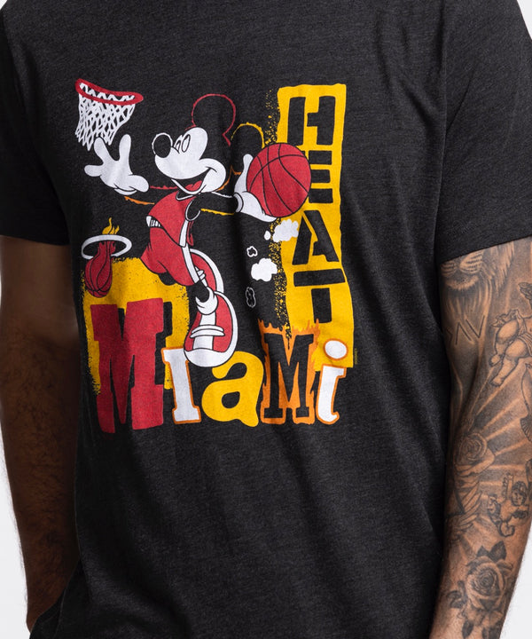 A man stands against a plain white background wearing blue jeans and a Homage Miami HEAT Mickey Basketball Tee by Homage, featuring a graphic of Mickey Mouse playing basketball and “Miami Heat” text.