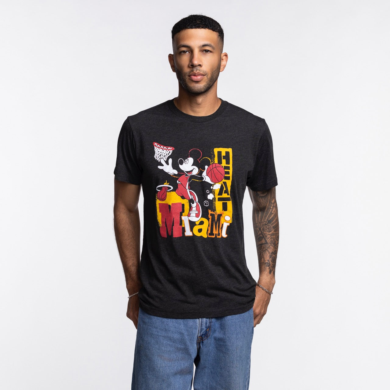 A man stands against a plain white background wearing blue jeans and a Homage Miami HEAT Mickey Basketball Tee by Homage, featuring a graphic of Mickey Mouse playing basketball and “Miami Heat” text.