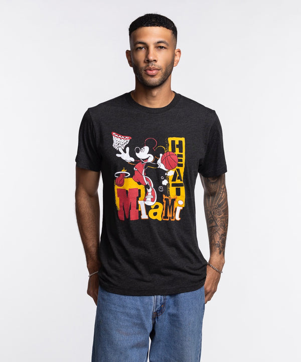 A man stands against a plain white background wearing blue jeans and a Homage Miami HEAT Mickey Basketball Tee by Homage, featuring a graphic of Mickey Mouse playing basketball and “Miami Heat” text.