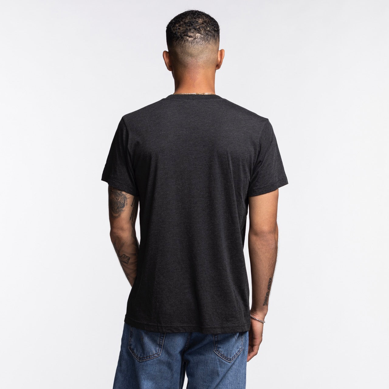 A person with short hair, wearing the Homage Miami HEAT Mickey Basketball Tee by Homage and blue jeans, stands facing away from the camera against a plain white background. The t-shirt is crafted from a soft polyester-cotton blend for comfort.