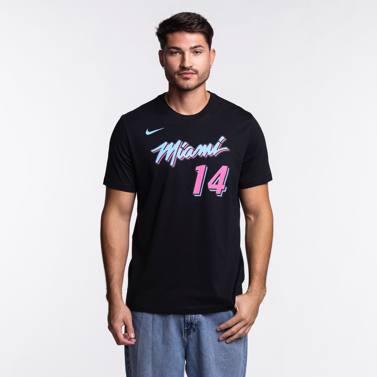 A man faces forward with one hand in his jeans pocket, wearing a black Nike Tyler Herro Original Vice Nights Name & Number Tee with pink and blue lettering, against a plain white background.