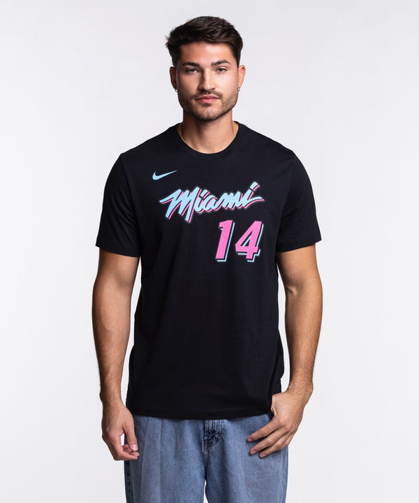 A man faces forward with one hand in his jeans pocket, wearing a black Nike Tyler Herro Original Vice Nights Name & Number Tee with pink and blue lettering, against a plain white background.