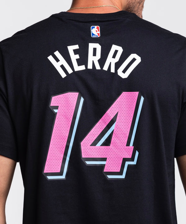 A man faces forward with one hand in his jeans pocket, wearing a black Nike Tyler Herro Original Vice Nights Name & Number Tee with pink and blue lettering, against a plain white background.