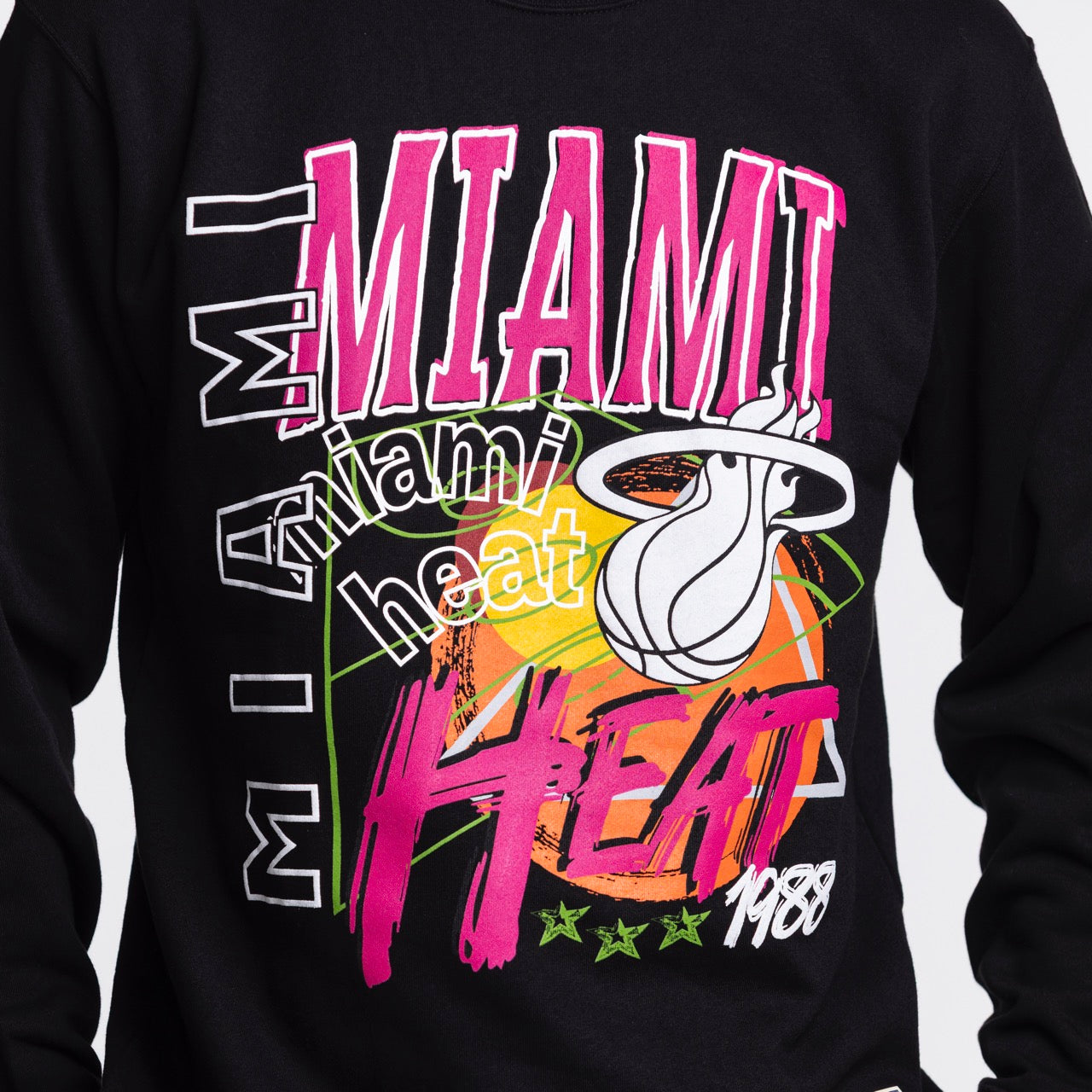 A person wearing the Mitchell & Ness Miami HEAT Original Vice Nights Crewneck, a black cotton-polyester sweatshirt featuring bold pink, white, and green MIAMI HEAT lettering, a basketball graphic, the team logo; standard fit; machine washable.
