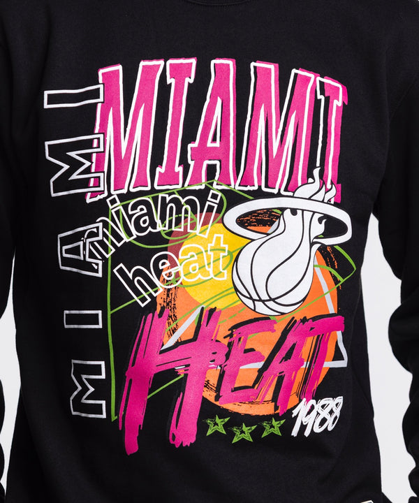 A man models the Mitchell & Ness Miami HEAT Original Vice Nights Crewneck—a black sweatshirt with colorful graphics in a standard fit—paired with blue jeans, standing against a white background and looking slightly to his left.
