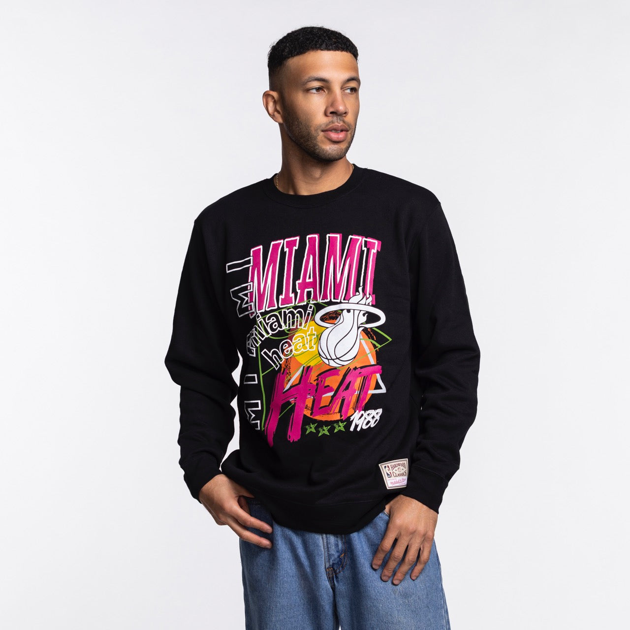 A man models the Mitchell & Ness Miami HEAT Original Vice Nights Crewneck—a black sweatshirt with colorful graphics in a standard fit—paired with blue jeans, standing against a white background and looking slightly to his left.