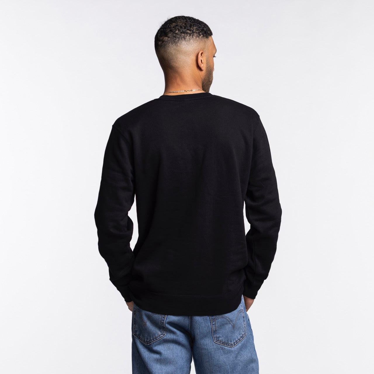 A man faces away from the camera, wearing the Mitchell & Ness Miami HEAT Original Vice Nights Crewneck in black with blue jeans, against a plain white background.