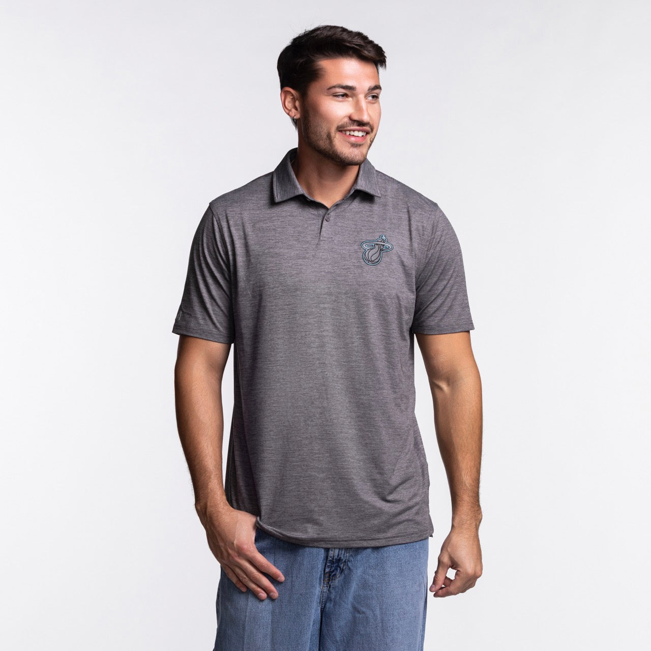 A man with short dark hair and a beard wears the Antigua Miami HEAT Original Vice Nights Polo by Antigua— a standard fit gray short-sleeve polo with a small chest logo—paired with blue jeans, standing and smiling against a plain light background.
