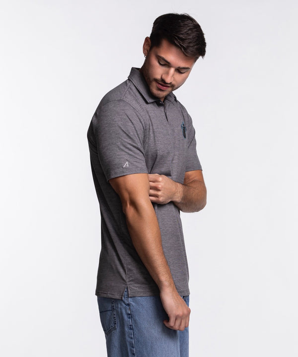A man with short dark hair and a beard wears the Antigua Miami HEAT Original Vice Nights Polo by Antigua— a standard fit gray short-sleeve polo with a small chest logo—paired with blue jeans, standing and smiling against a plain light background.