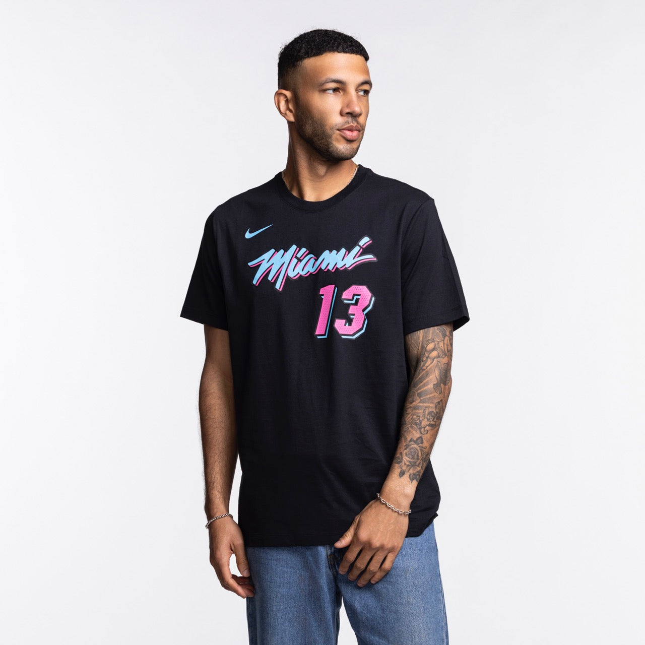 A man with short hair and a beard wears the Bam Adebayo Nike Original Vice Nights Name & Number Tee, featuring black washable cotton, pink and blue Miami basketball graphics, and number 13. He has a left forearm tattoo and stands against a white background.
