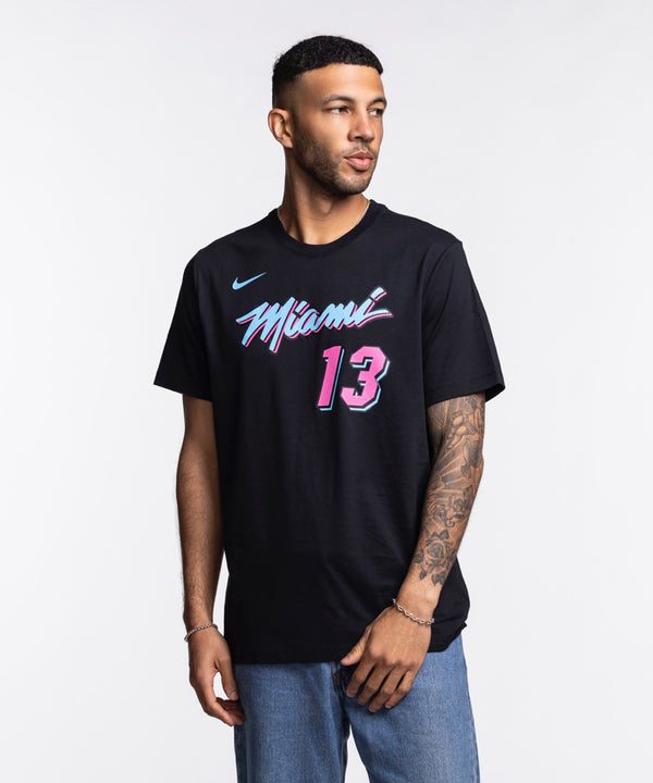 A man with short hair and a beard wears the Bam Adebayo Nike Original Vice Nights Name & Number Tee, featuring black washable cotton, pink and blue Miami basketball graphics, and number 13. He has a left forearm tattoo and stands against a white background.