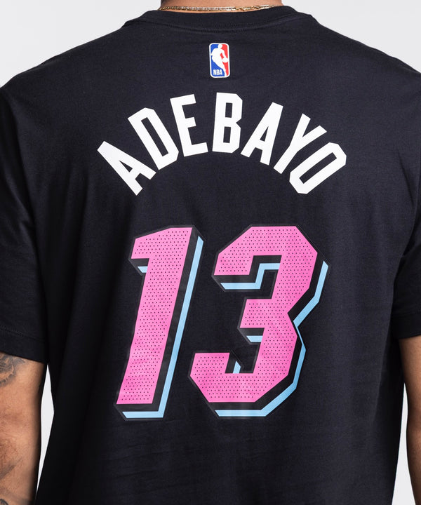A man with short hair and a beard wears the Bam Adebayo Nike Original Vice Nights Name & Number Tee, featuring black washable cotton, pink and blue Miami basketball graphics, and number 13. He has a left forearm tattoo and stands against a white background.