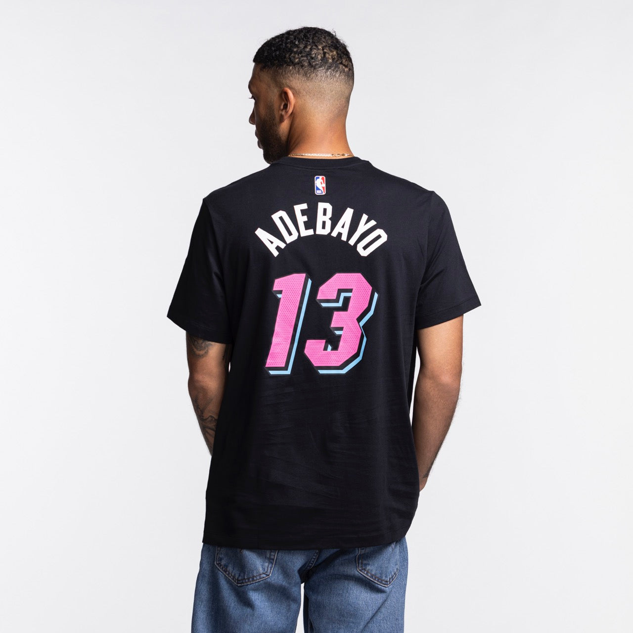 A man faces away wearing the Nike Bam Adebayo Original Vice Nights Name & Number Tee, a black cotton basketball-style shirt with ADEBAYO and 13 in pink and blue. He pairs it with blue jeans against a light background. Machine washable for easy care.