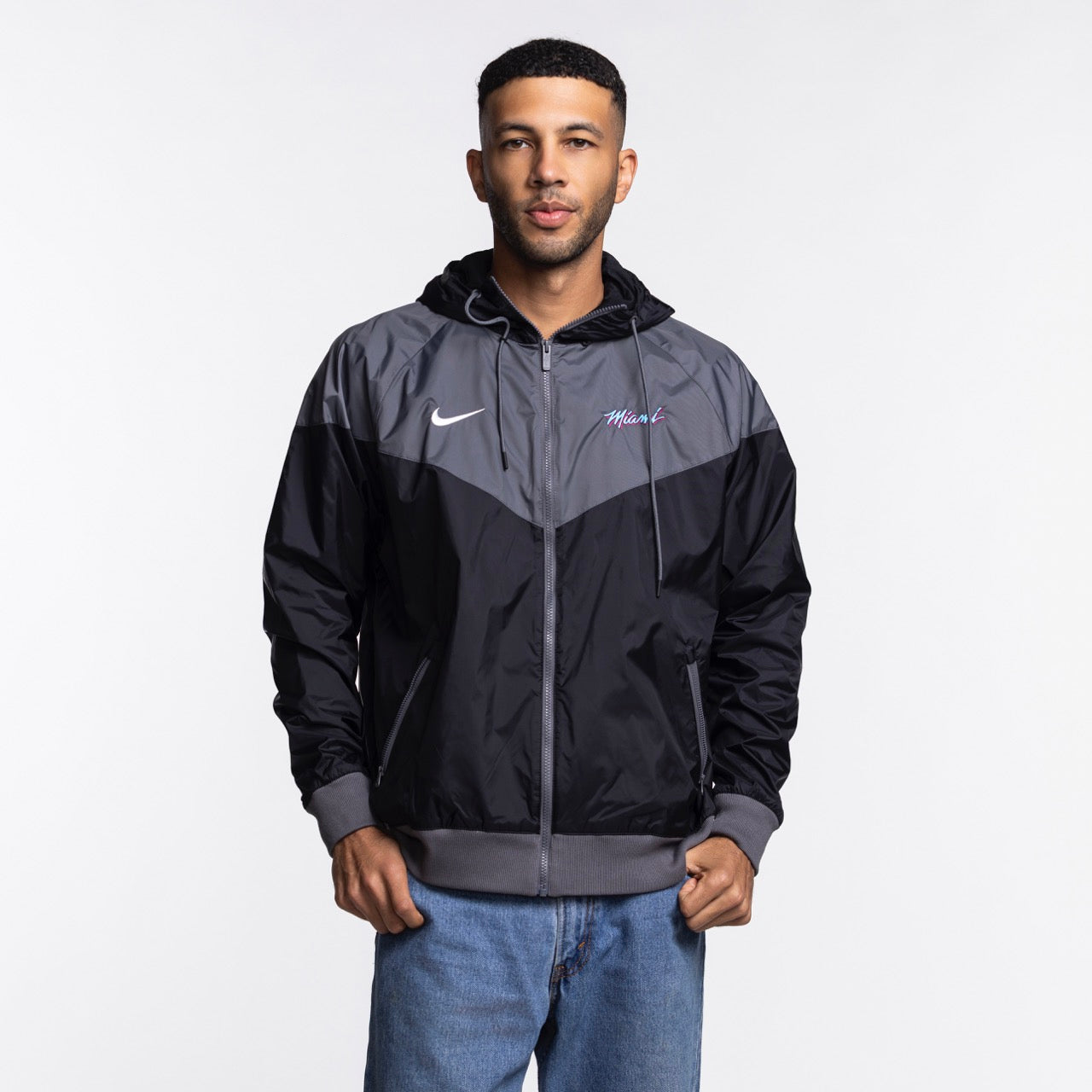A man wears the Nike Miami HEAT Original Vice Nights Windbreaker Jacket, a black and gray standard-fit polyester hoodie, paired with light blue jeans, standing before a plain white background.