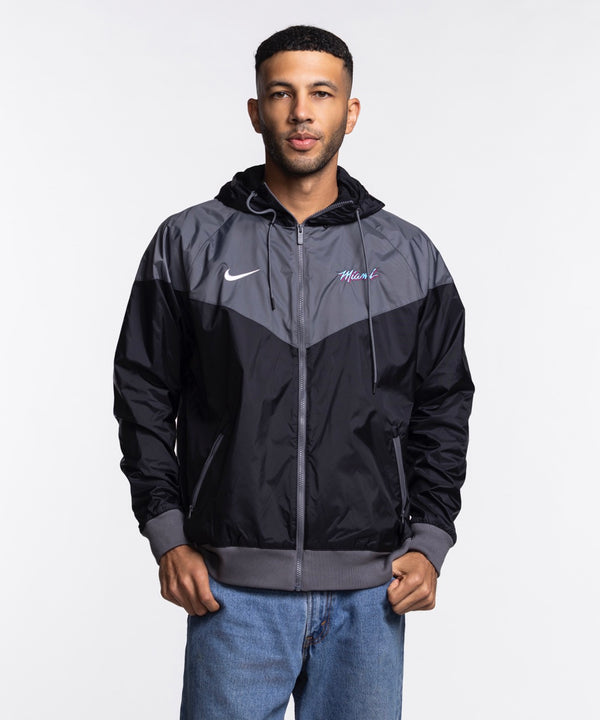 A man wears the Nike Miami HEAT Original Vice Nights Windbreaker Jacket, a black and gray standard-fit polyester hoodie, paired with light blue jeans, standing before a plain white background.