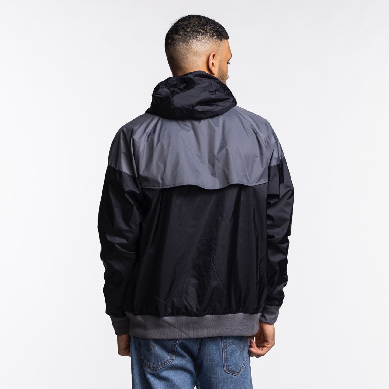 A man with short hair stands with his back to the camera, wearing a Nike Miami HEAT Original Vice Nights Windbreaker Jacket and blue jeans against a plain white background.