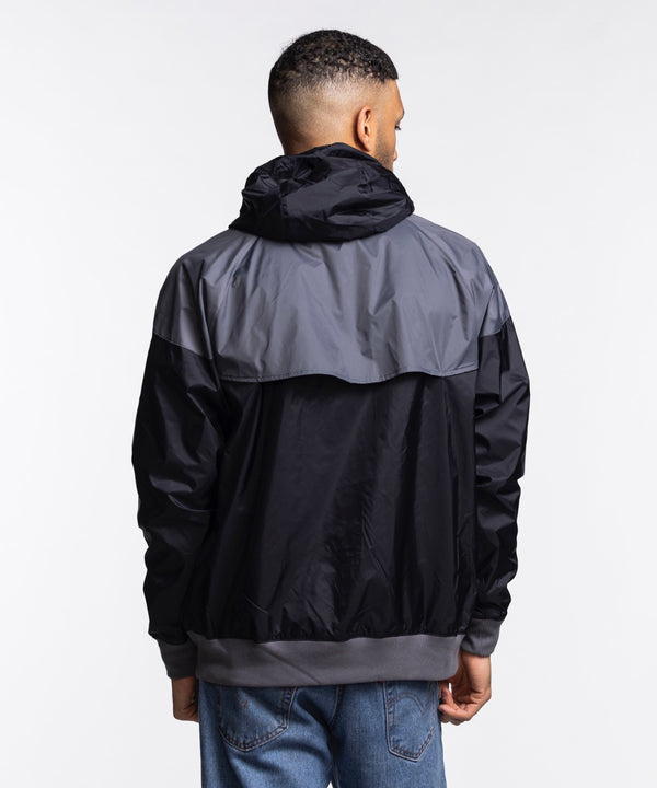 A man wears the Nike Miami HEAT Original Vice Nights Windbreaker Jacket, a black and gray standard-fit polyester hoodie, paired with light blue jeans, standing before a plain white background.