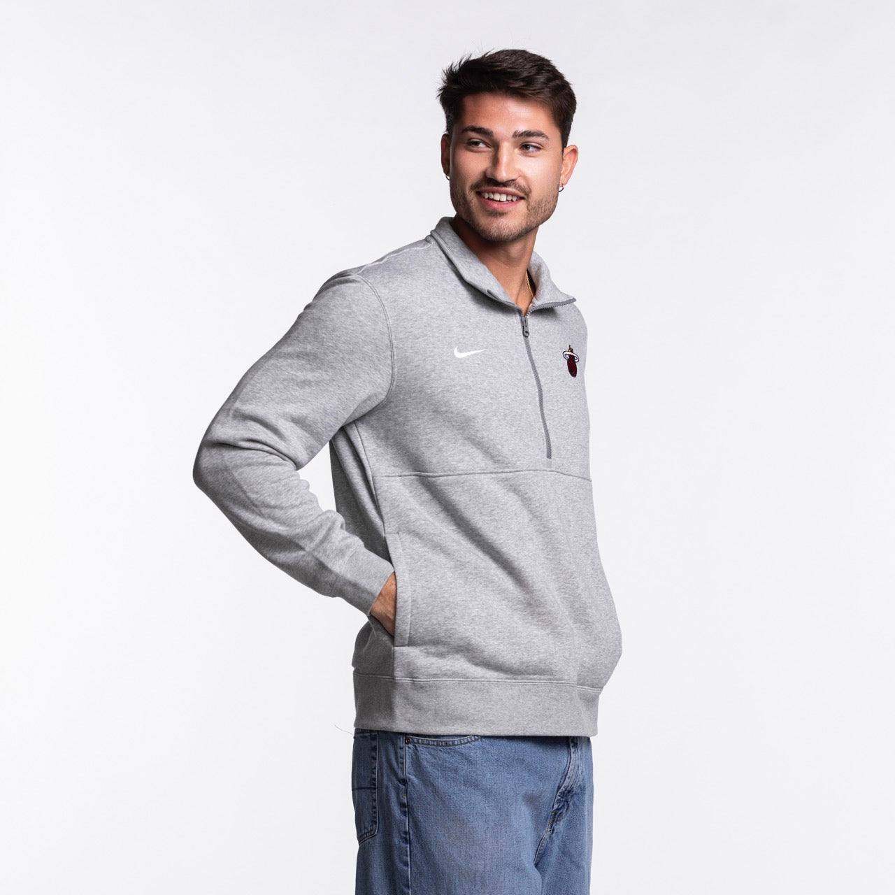 A man wears a Nike Miami HEAT Fleece 1/2 Zip Jacket and blue jeans, standing with his hands in his pockets and smiling to the side against a plain white background.