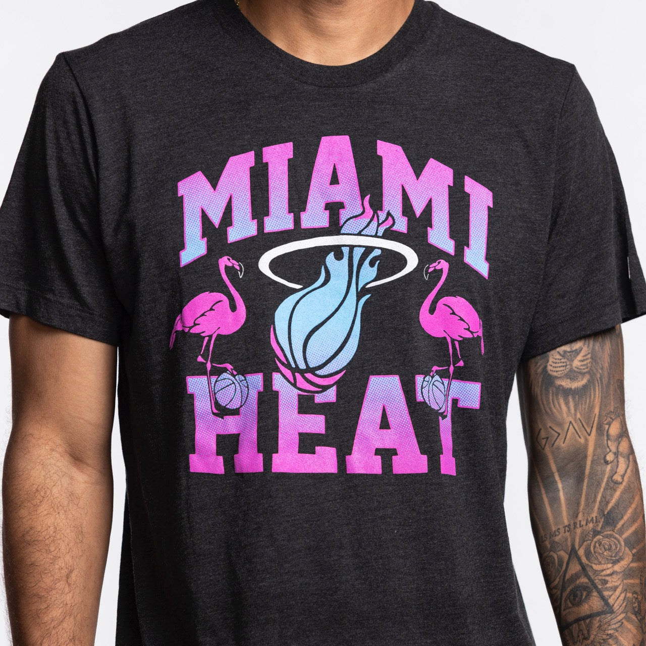 A person wears the Homage Miami HEAT Original Vice Nights Flamingo Tee by Homage, featuring pink and blue lettering, two flamingos with basketballs, and a flaming basketball logo. The soft poly-cotton-rayon blend fits standard; part of their tattooed arm is visible.