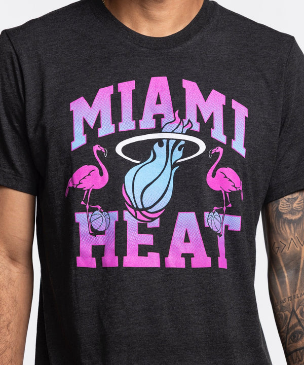 A man wears the Homage Miami HEAT Original Vice Nights Flamingo Tee, a standard fit black shirt with a colorful flamingos and basketball logo. Made of soft polyester-cotton-rayon by Homage, it’s machine washable cold. He stands against a plain white background.