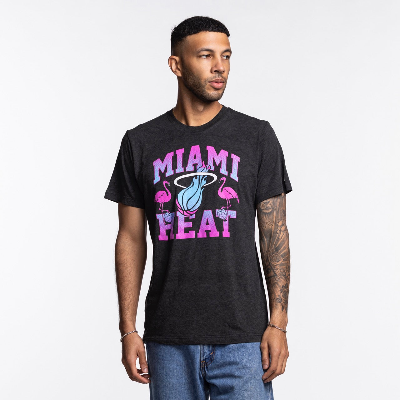 A man wears the Homage Miami HEAT Original Vice Nights Flamingo Tee, a standard fit black shirt with a colorful flamingos and basketball logo. Made of soft polyester-cotton-rayon by Homage, it’s machine washable cold. He stands against a plain white background.