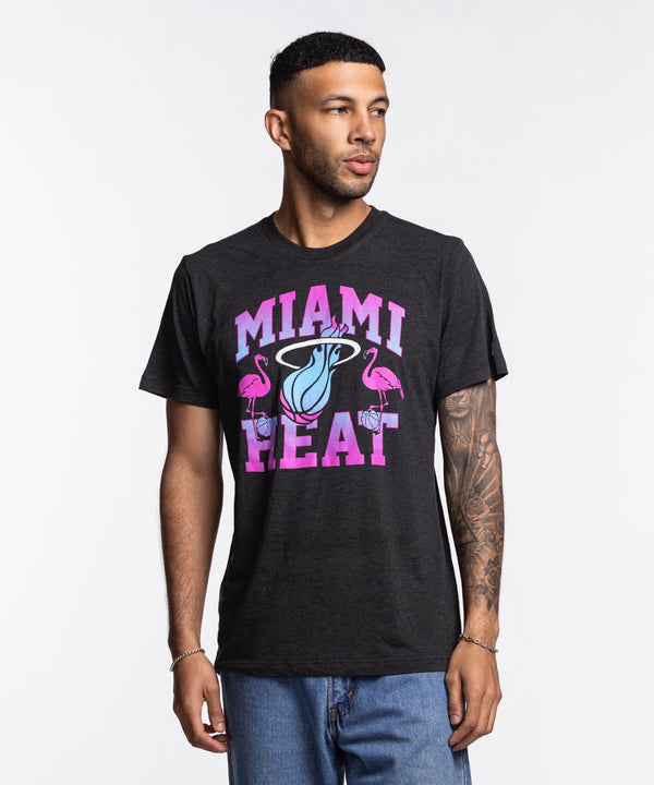 A man wears the Homage Miami HEAT Original Vice Nights Flamingo Tee, a standard fit black shirt with a colorful flamingos and basketball logo. Made of soft polyester-cotton-rayon by Homage, it’s machine washable cold. He stands against a plain white background.