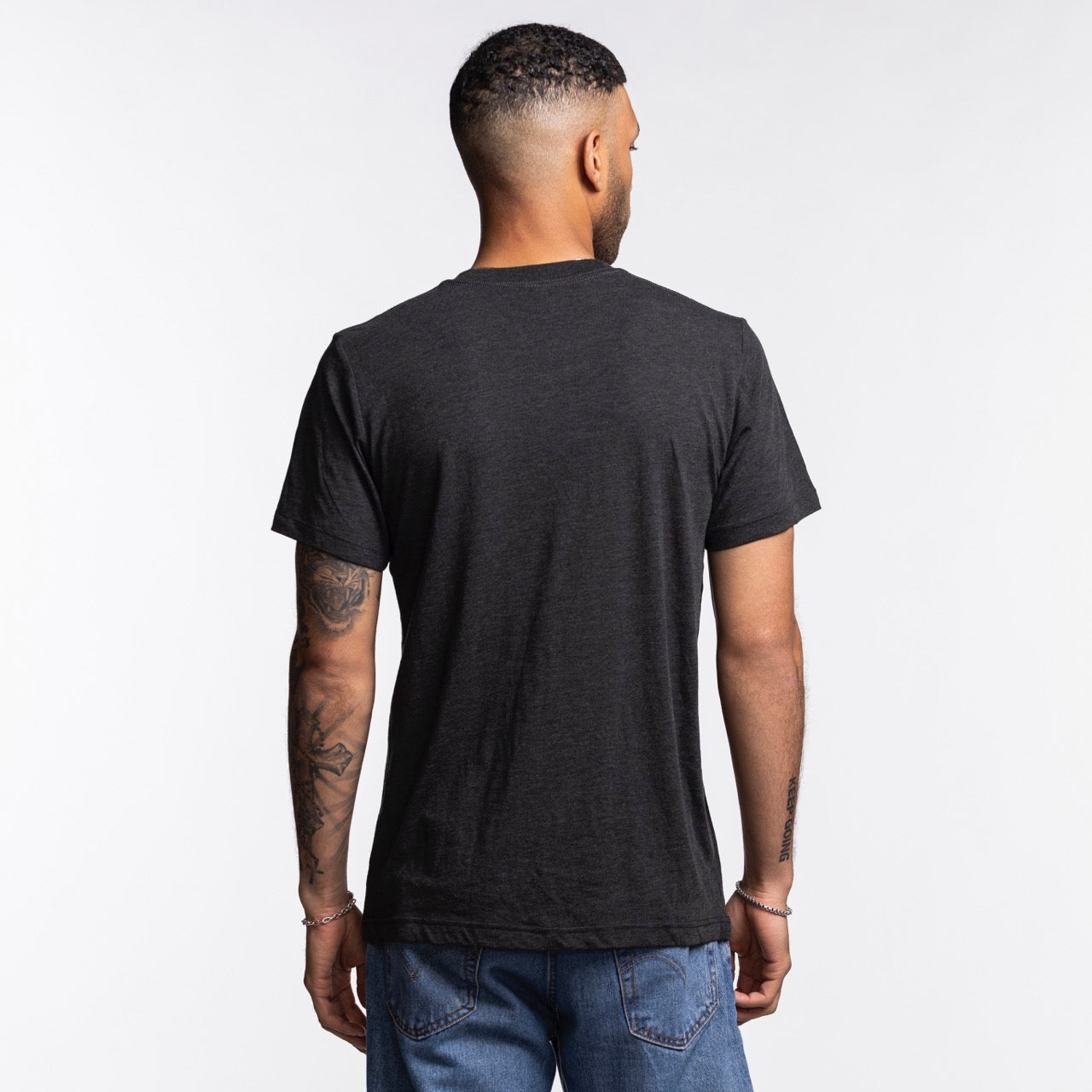 A man with short hair faces away from the camera, showing his tattooed right arm and the back of a Homage Miami HEAT Original Vice Nights Flamingo Tee in a standard fit, paired with blue jeans against a plain light background.