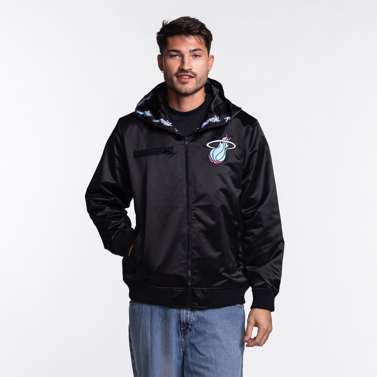 A man in a black Mitchell & Ness Miami HEAT Original Vice Nights Satin Bomber with a hood, made from polyester in a standard fit, pairs it with light blue jeans. He stands facing forward against a white background. Machine wash cold.
