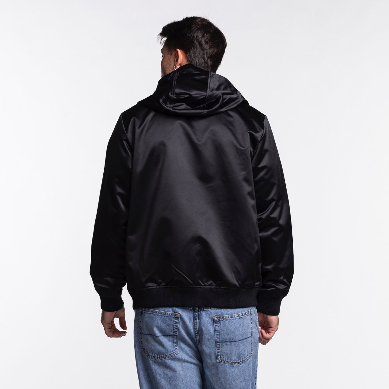 A man wearing a Mitchell & Ness Miami HEAT Original Vice Nights Satin Bomber in shiny black stands with his back to the camera, paired with light blue jeans, against a plain white background.