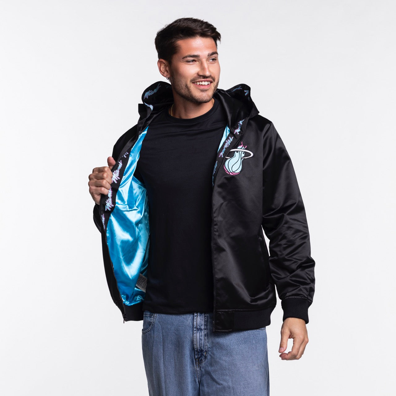 A man wearing the Mitchell & Ness Miami HEAT Original Vice Nights Satin Bomber stands against a white background, holding one side of the jacket open and smiling, paired with a black tee and blue jeans.