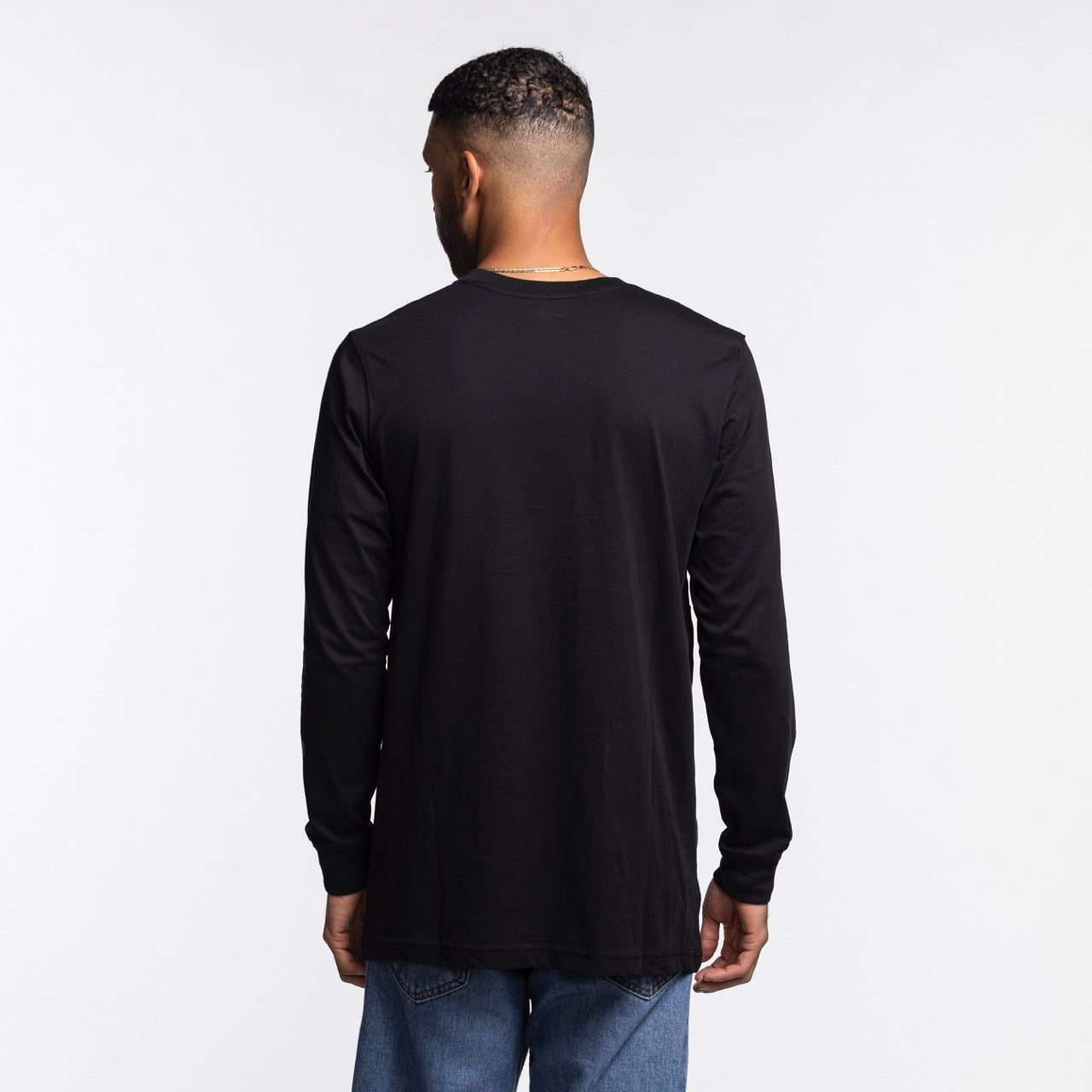 A man with short dark hair stands with his back to the camera, wearing a New Era Miami HEAT Original Vice Nights Wordmark Long Sleeve Tee and blue jeans against a plain white background.