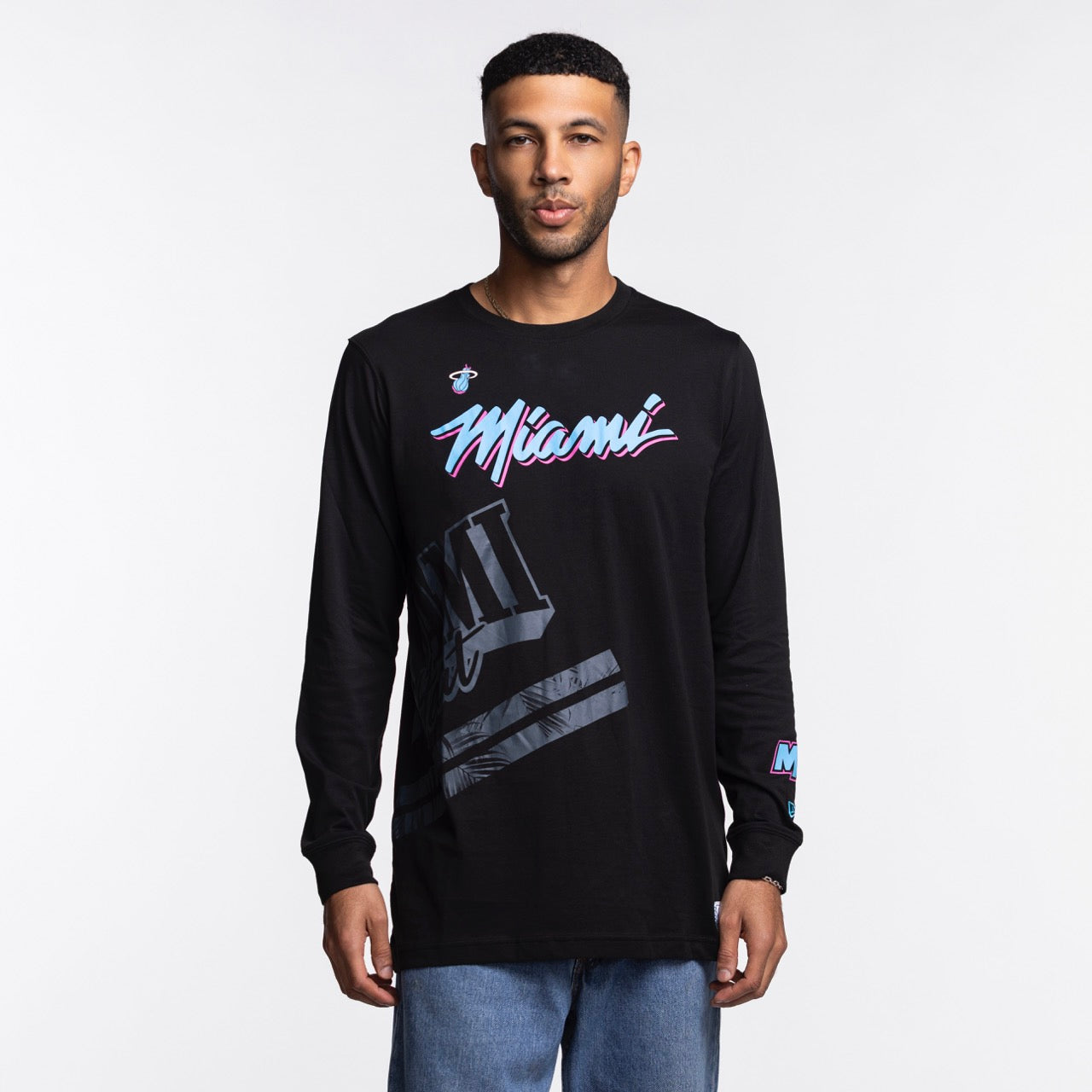 A man stands facing forward against a plain white background wearing a New Era Miami HEAT Original Vice Nights Wordmark Long Sleeve Tee, a black 100% cotton shirt featuring colorful Miami text and graphic designs on the front and sleeve.