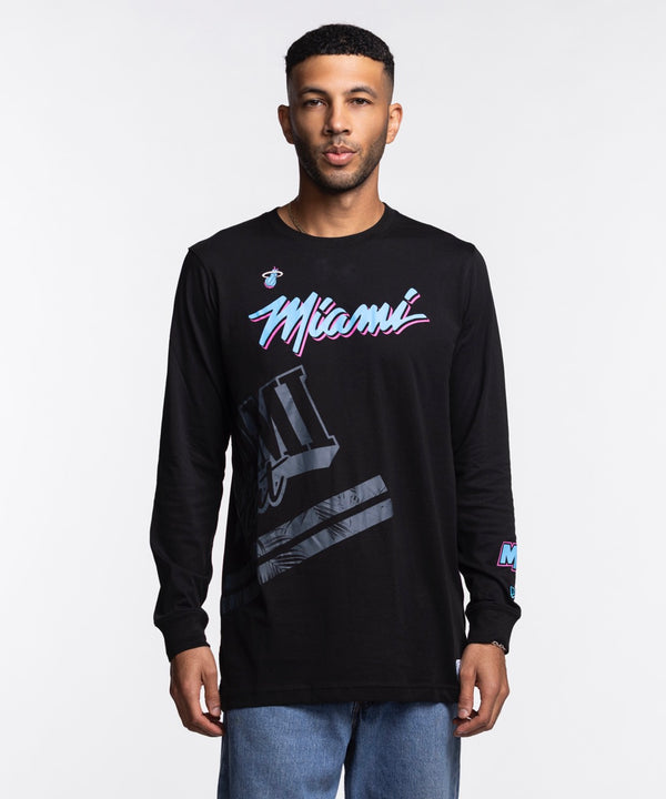 A man stands facing forward against a plain white background wearing a New Era Miami HEAT Original Vice Nights Wordmark Long Sleeve Tee, a black 100% cotton shirt featuring colorful Miami text and graphic designs on the front and sleeve.