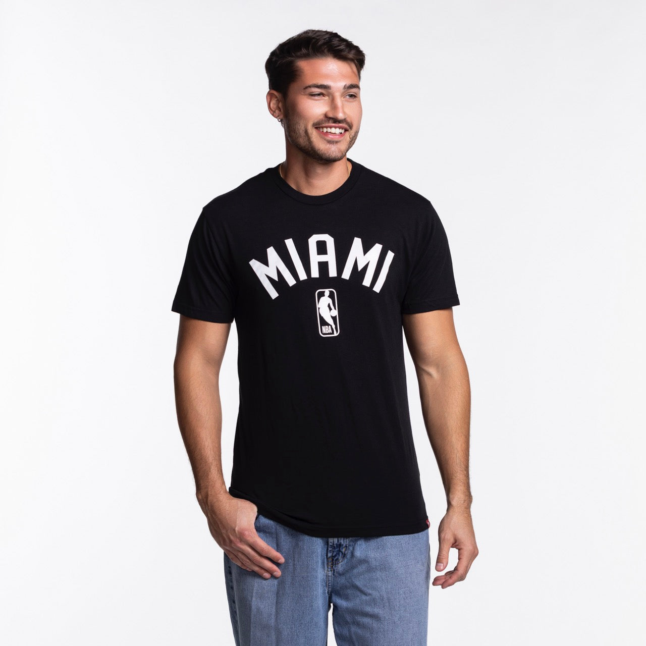 A man with short dark hair and a beard is wearing the Sportiqe Miami HEAT NBA Tee by Sportiqe, featuring MIAMI and a basketball logo, paired with blue jeans, standing and smiling against a plain white background.