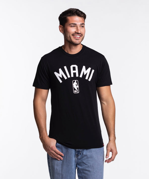 A man with short dark hair and a beard is wearing the Sportiqe Miami HEAT NBA Tee by Sportiqe, featuring MIAMI and a basketball logo, paired with blue jeans, standing and smiling against a plain white background.