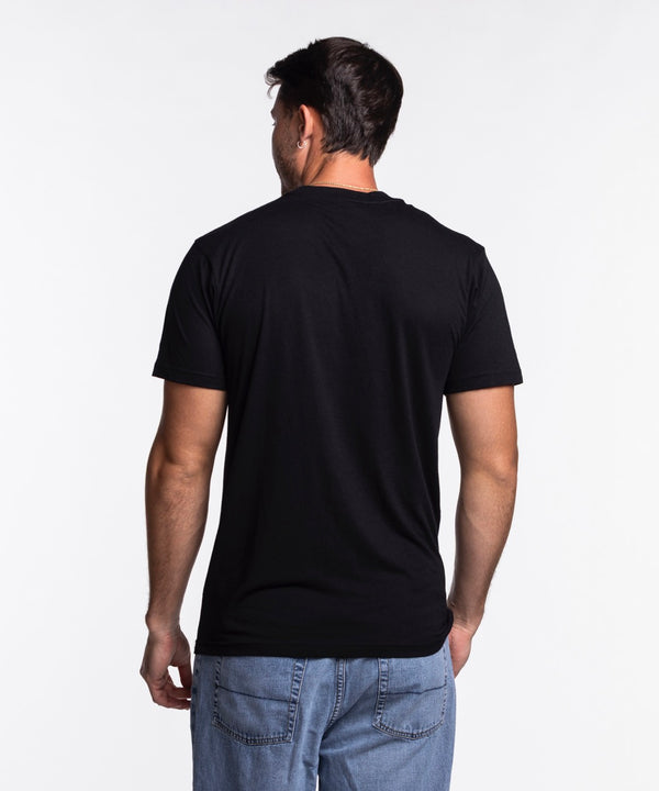 A man with short dark hair and a beard is wearing the Sportiqe Miami HEAT NBA Tee by Sportiqe, featuring MIAMI and a basketball logo, paired with blue jeans, standing and smiling against a plain white background.