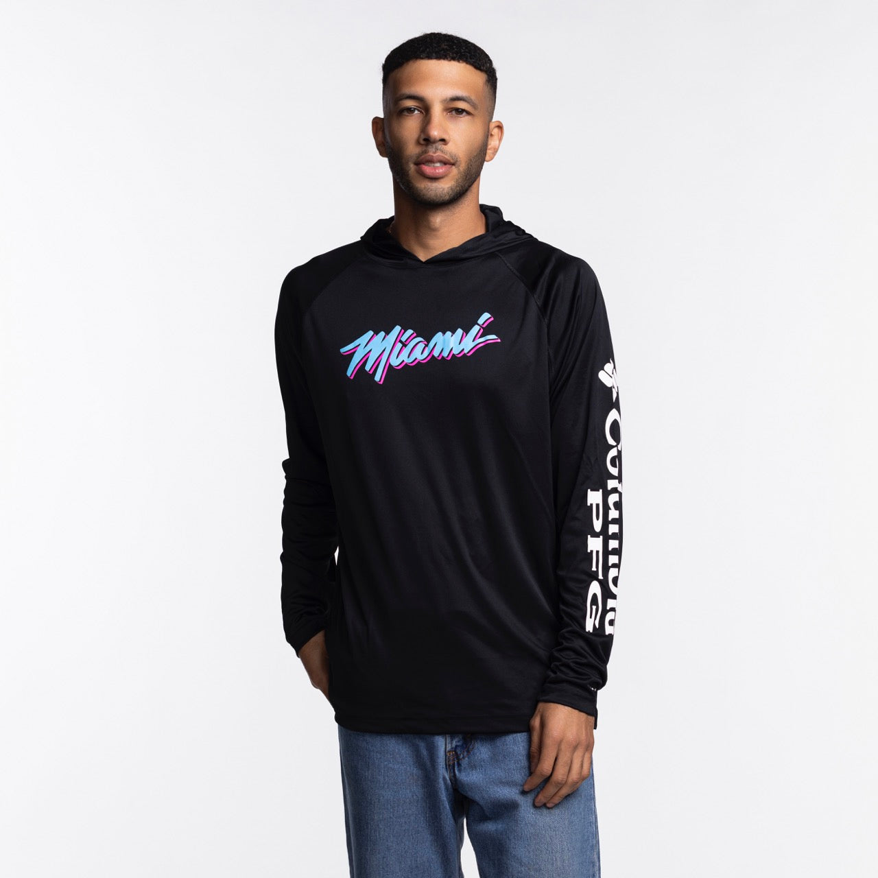 A man stands against a white background wearing the Columbia Miami HEAT Original Vice Nights Long Sleeve Hoodie Tee by Columbia Outdoor in black, featuring colorful 