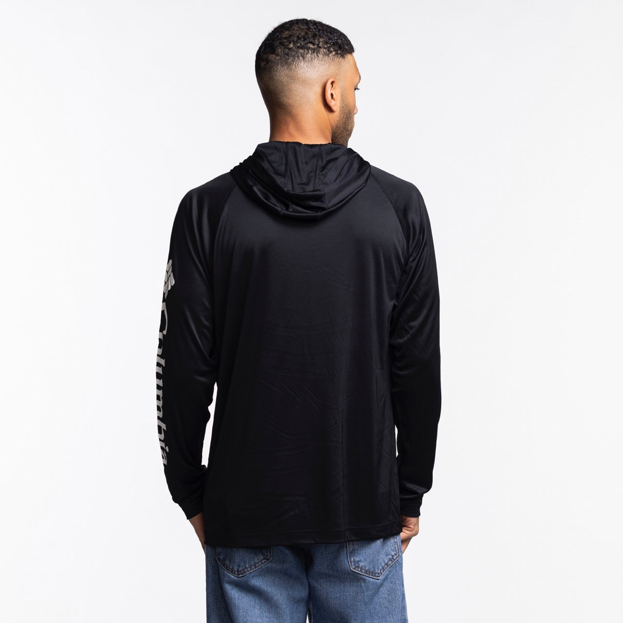 A man with short hair, seen from behind, wears a Columbia Miami HEAT Original Vice Nights Long Sleeve Hoodie Tee by Columbia Outdoor and blue jeans, standing against a plain white background.